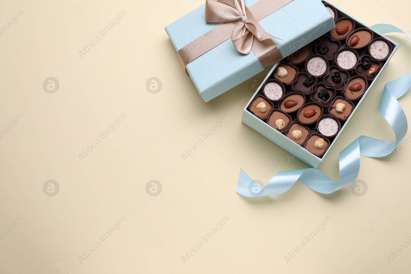 Photo of Open box of delicious chocolate candies and light blue ribbon on beige background, flat lay. Space for text