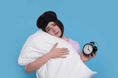 Unhappy young woman with sleep mask, alarm clock and pillow on light blue background. Insomnia problem