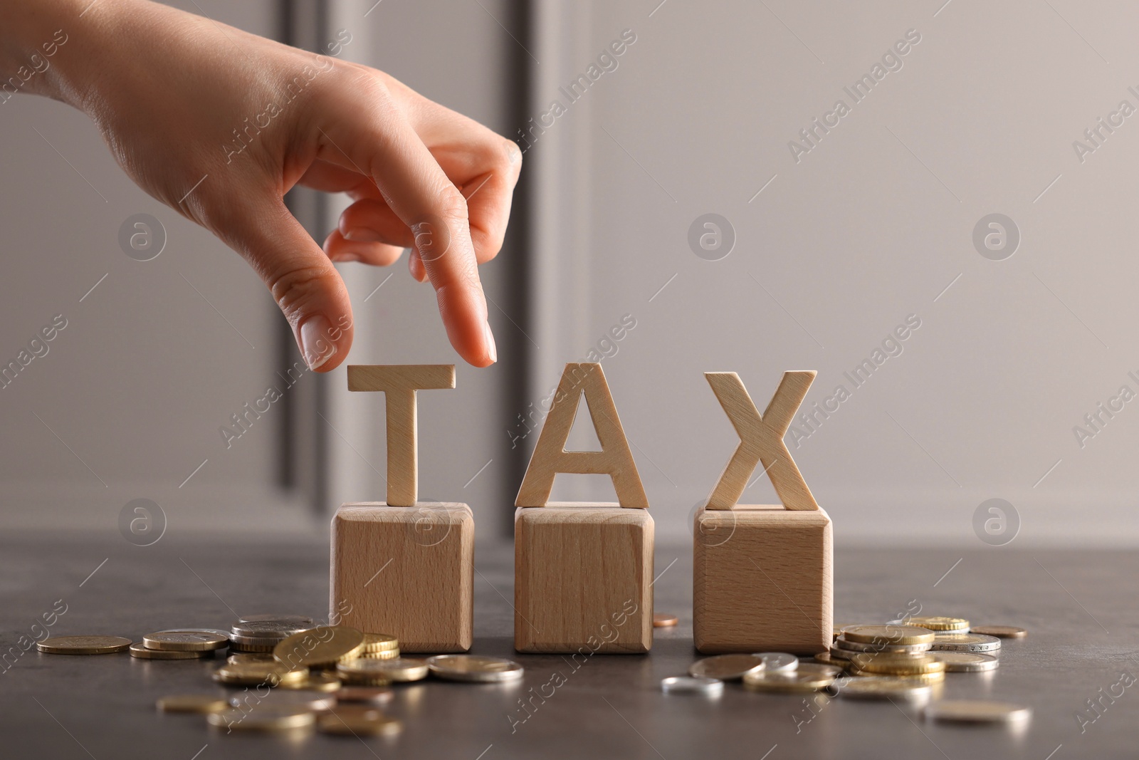 Photo of Woman with word Tax, wooden cubes and coins at grey table, closeup