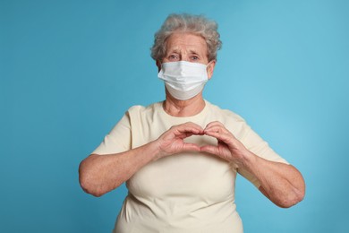 Photo of Elderly woman in medical mask making heart with her hands on light blue background