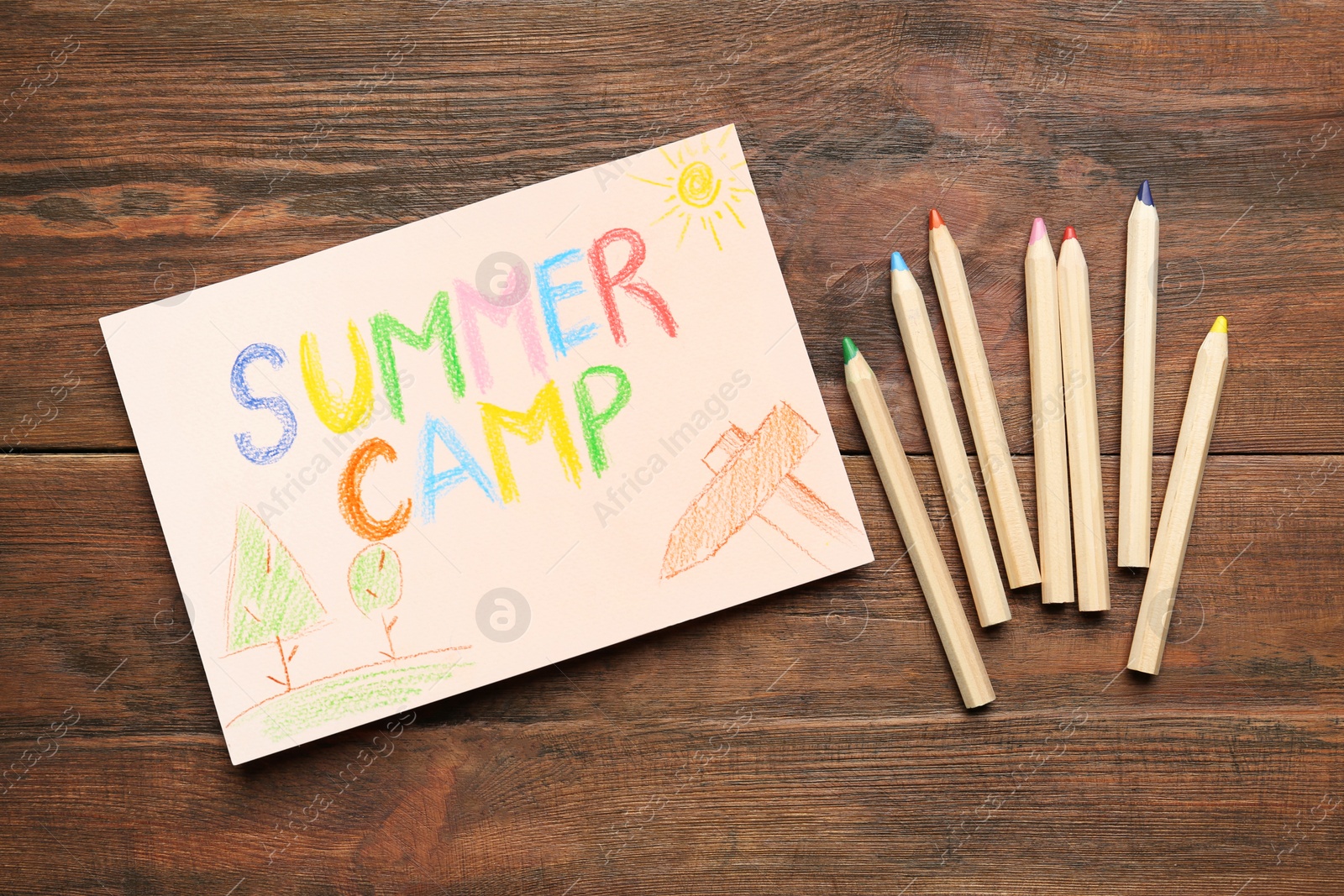 Photo of Card with text SUMMER CAMP, drawings and colorful pencils on wooden table, flat lay