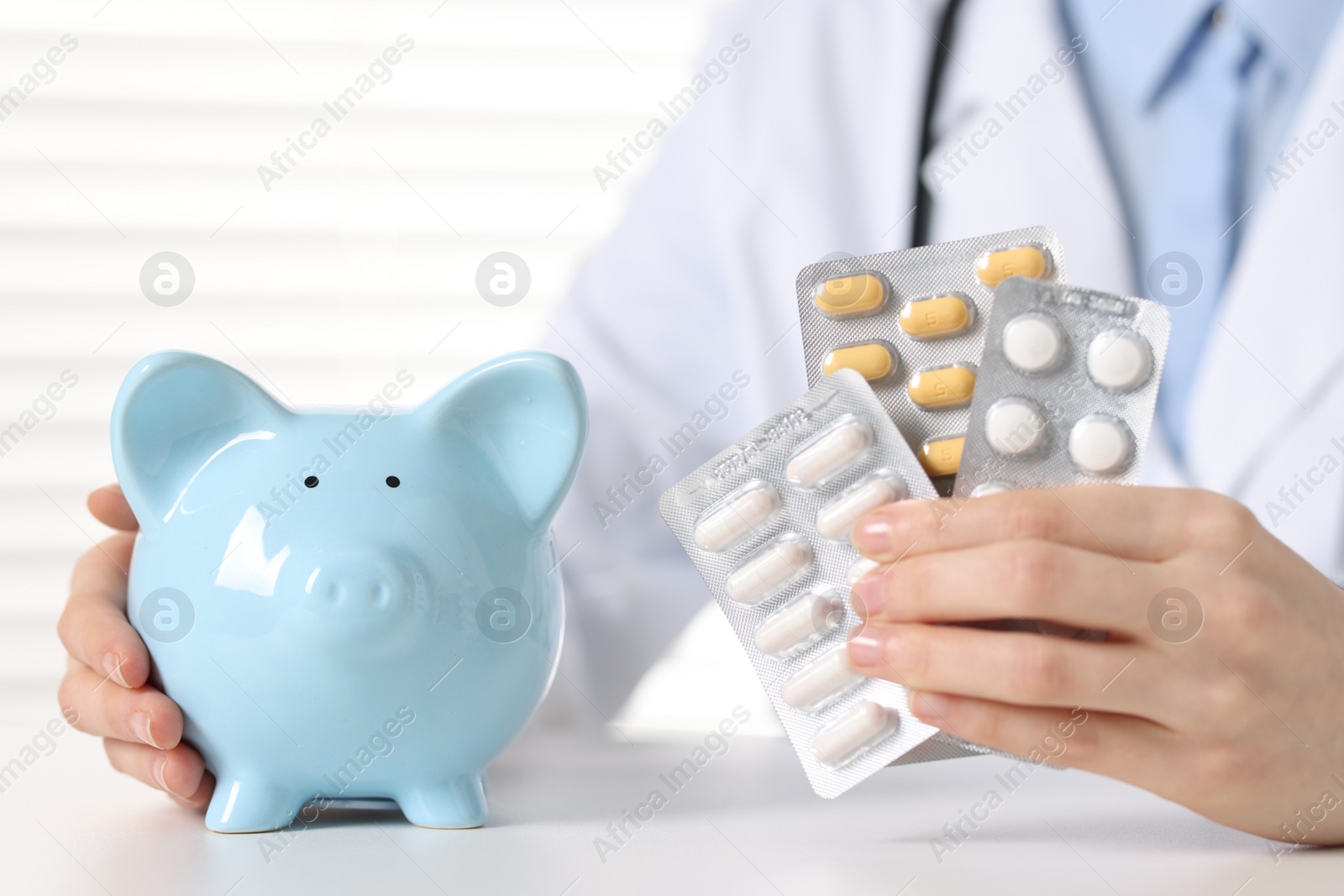 Photo of Doctor with pills and piggy bank at white table indoors, closeup