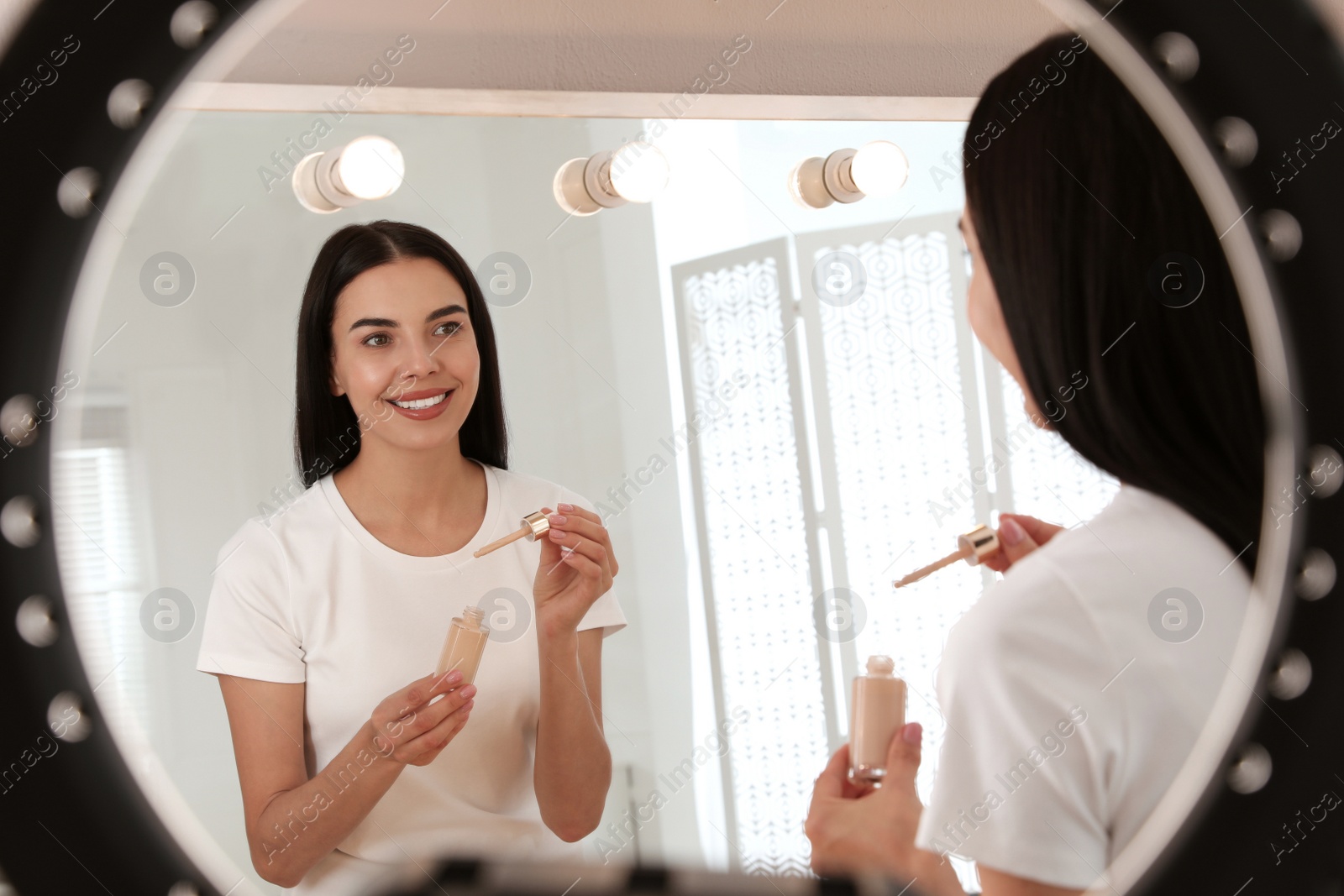 Photo of Beautiful young woman with liquid foundation near mirror indoors, view through ring lamp