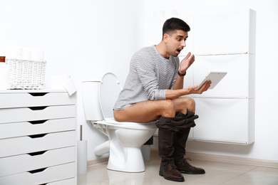 Photo of Emotional man with tablet sitting on toilet bowl in bathroom