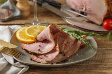 Photo of Slices of delicious baked ham, orange, rosemary, fork, and knife on wooden table, closeup
