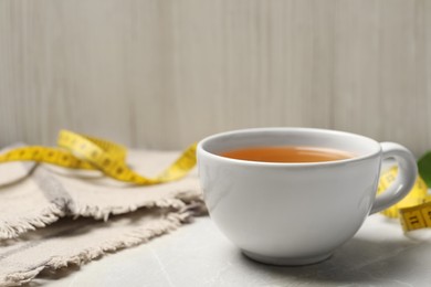 Cup of herbal diet tea and measuring tape on light marble table against wooden wall, space for text. Weight loss concept