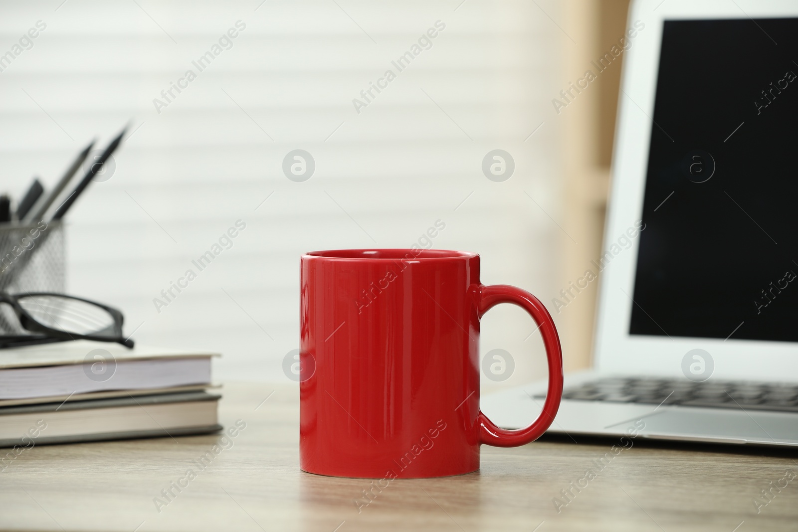 Photo of Red ceramic mug, notebooks and laptop on wooden table indoors