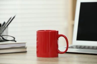 Photo of Red ceramic mug, notebooks and laptop on wooden table indoors