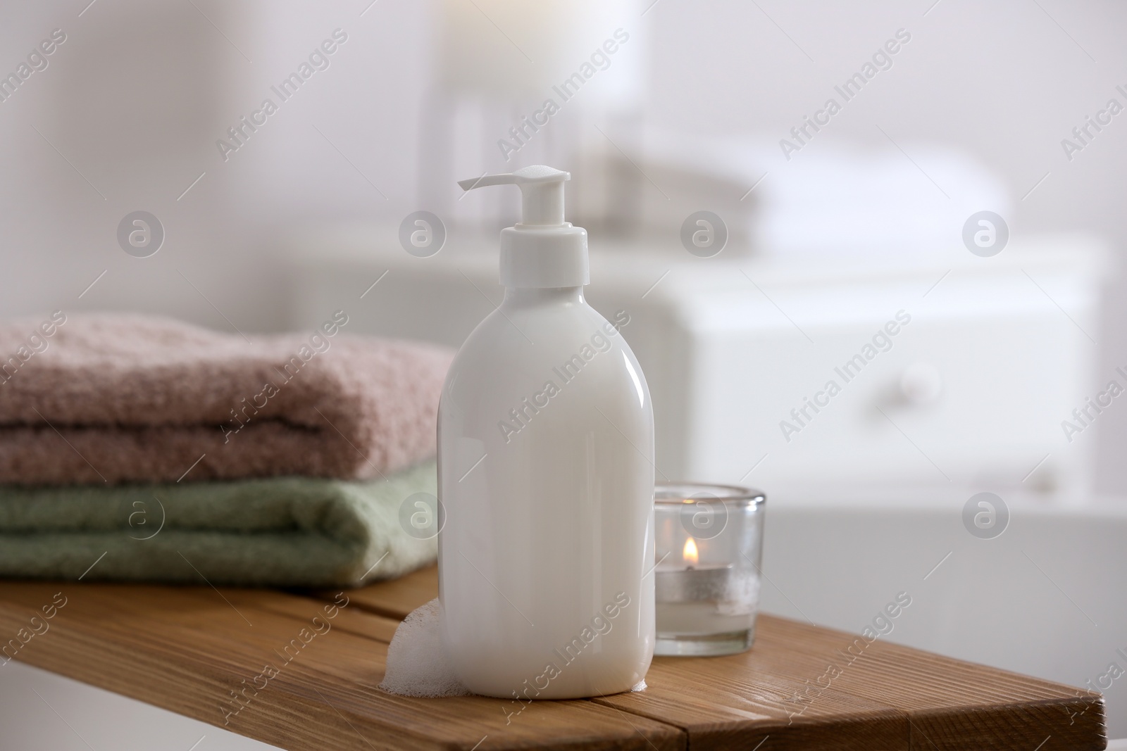 Photo of Bottle of bubble bath with foam, towels and candle on tub in bathroom