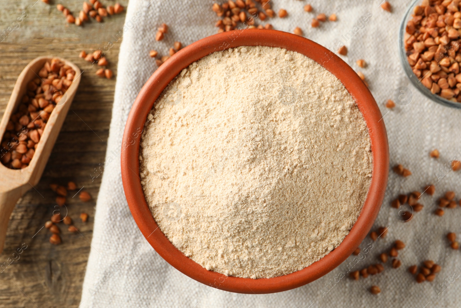 Photo of Bowl of buckwheat flour, scoop cloth on wooden table, flat lay