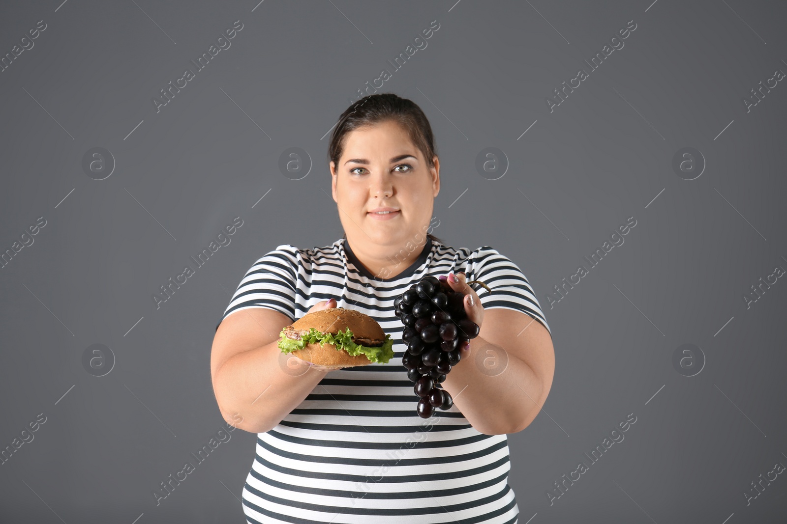 Photo of Overweight woman with grapes and hamburger on gray background