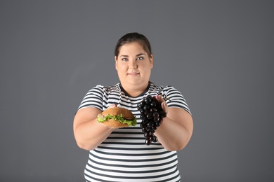 Overweight woman with grapes and hamburger on gray background