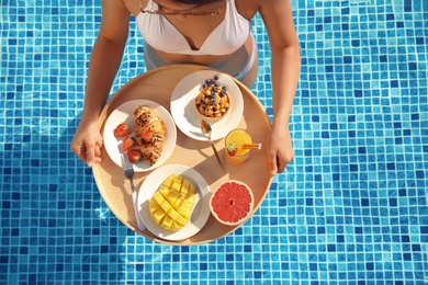 Young woman with delicious breakfast on floating tray in swimming pool, top view