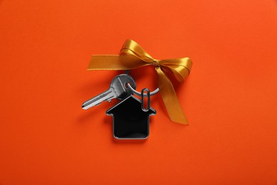 Photo of Key with trinket in shape of house and yellow bow on orange background, top view. Housewarming party