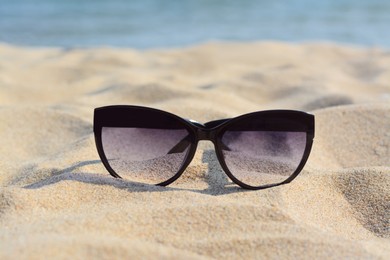 Stylish sunglasses on sandy beach near sea, closeup