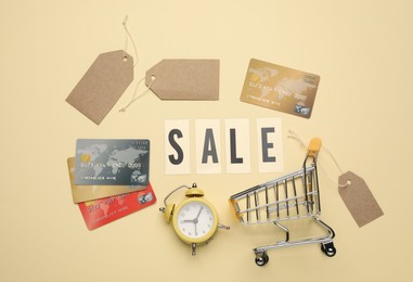Photo of Shopping cart, tags, credit cards, alarm clock and word Sale on beige background, flat lay