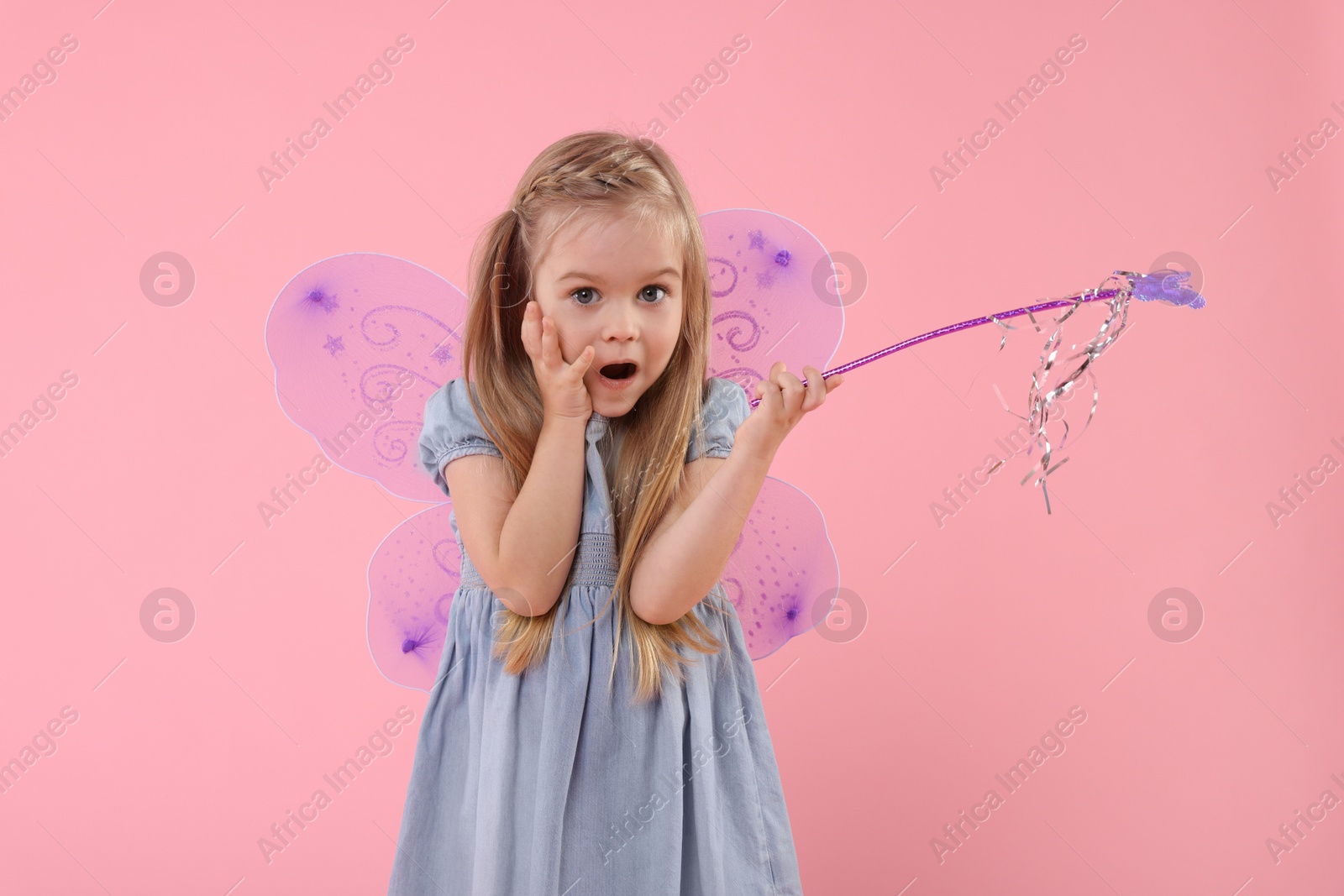 Photo of Surprised little girl in fairy costume with violet wings and magic wand on pink background