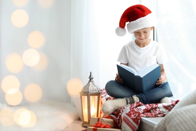 Cute little child in Santa hat reading Christmas story while sitting on windowsill at home