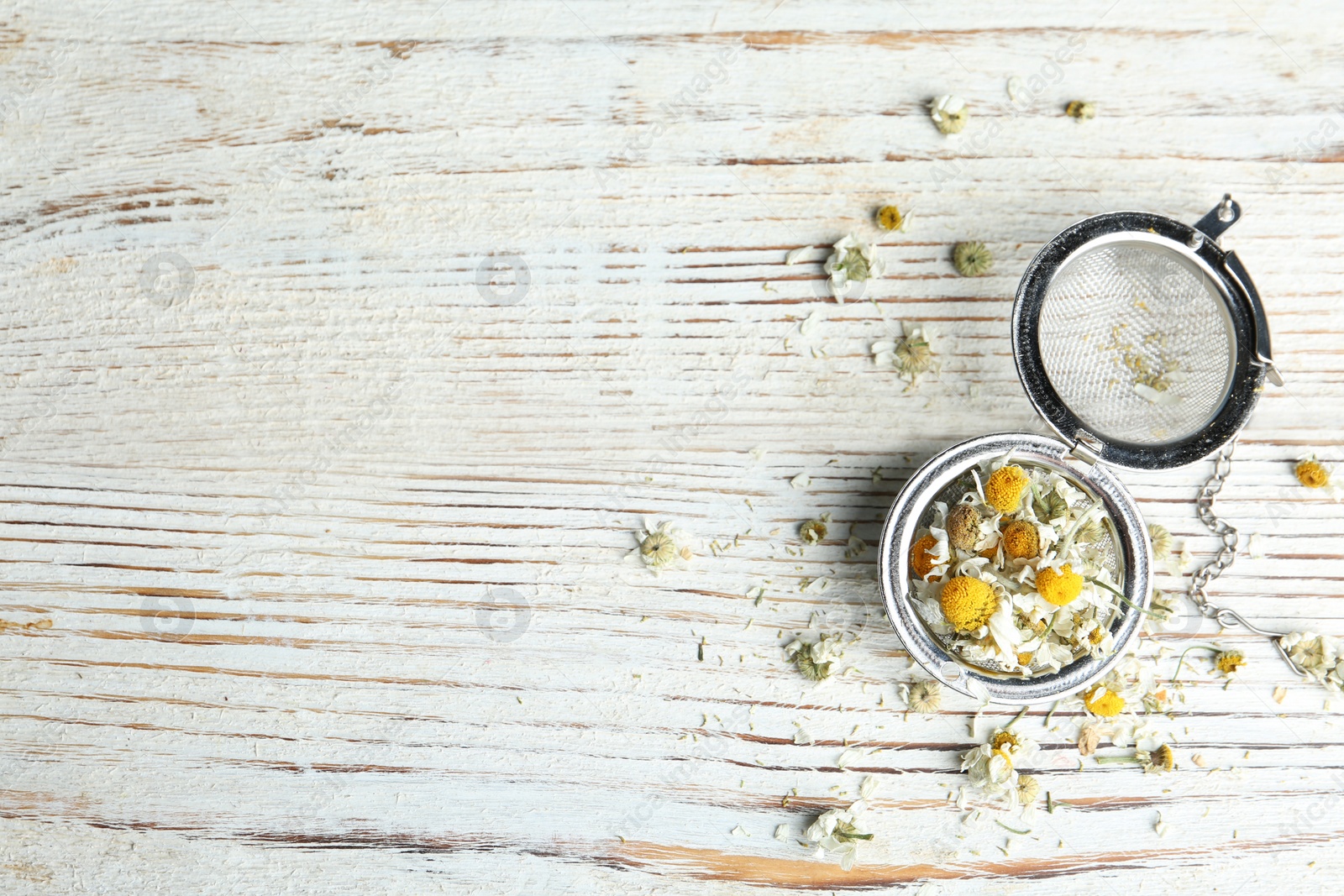 Photo of Dry chamomile flowers in infuser on white wooden table, flat lay. Space for text