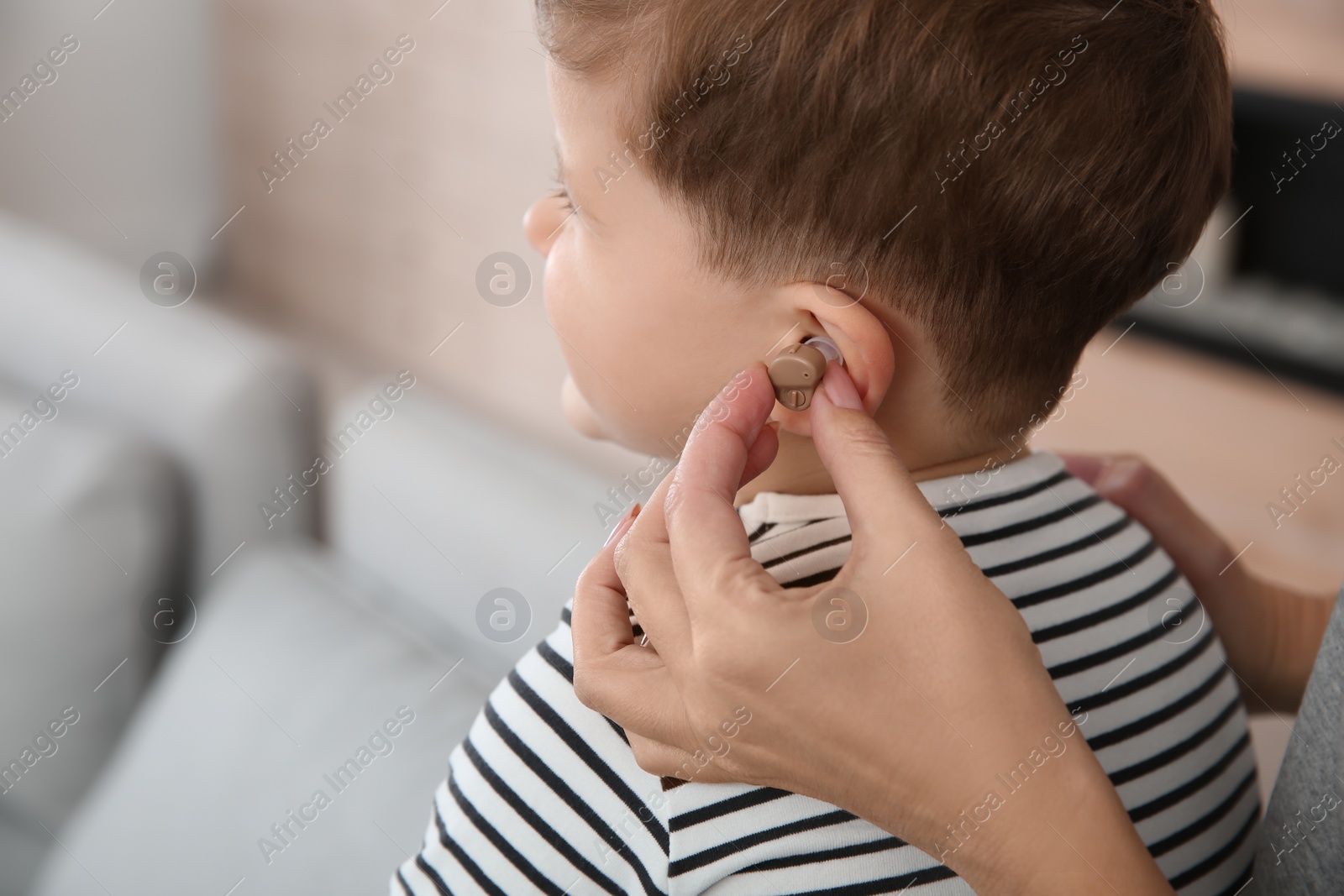 Photo of Young woman putting hearing aid in little son's ear indoors, closeup