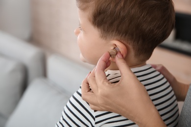 Young woman putting hearing aid in little son's ear indoors, closeup