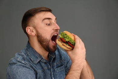 Young man eating tasty burger on grey background