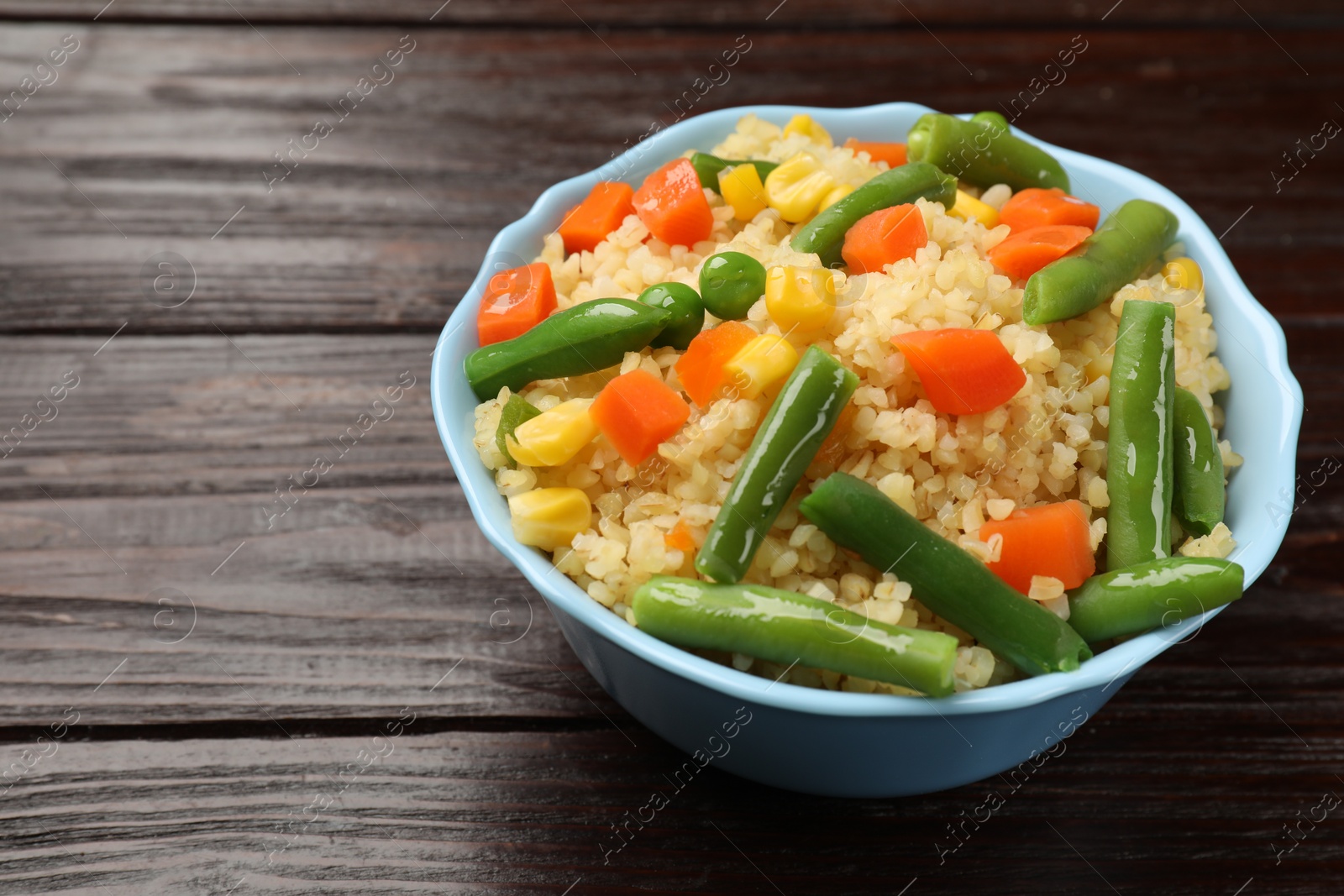 Photo of Delicious bulgur with vegetables in bowl on wooden table, closeup. Space for text