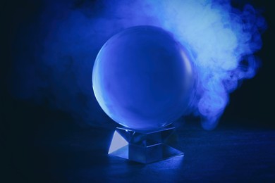 Photo of Magic crystal ball on table and smoke against dark background. Making predictions