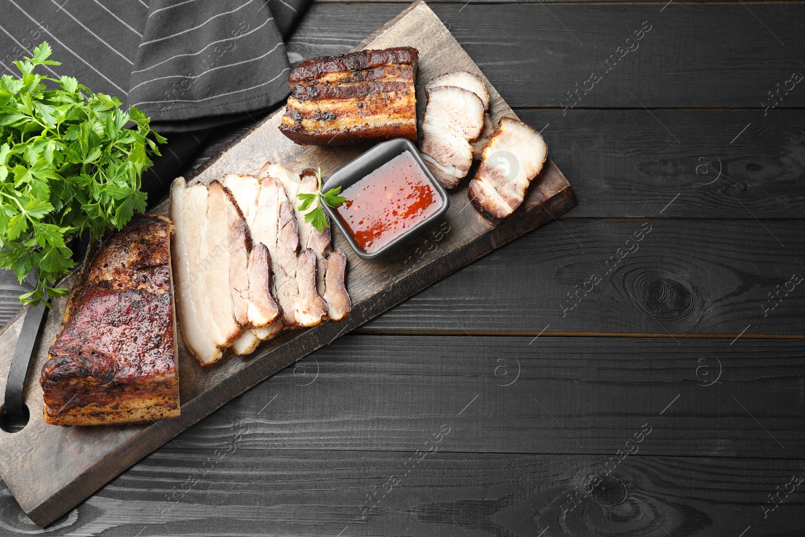 Photo of Pieces of baked pork belly served with sauce and parsley on black wooden table, top view. Space for text