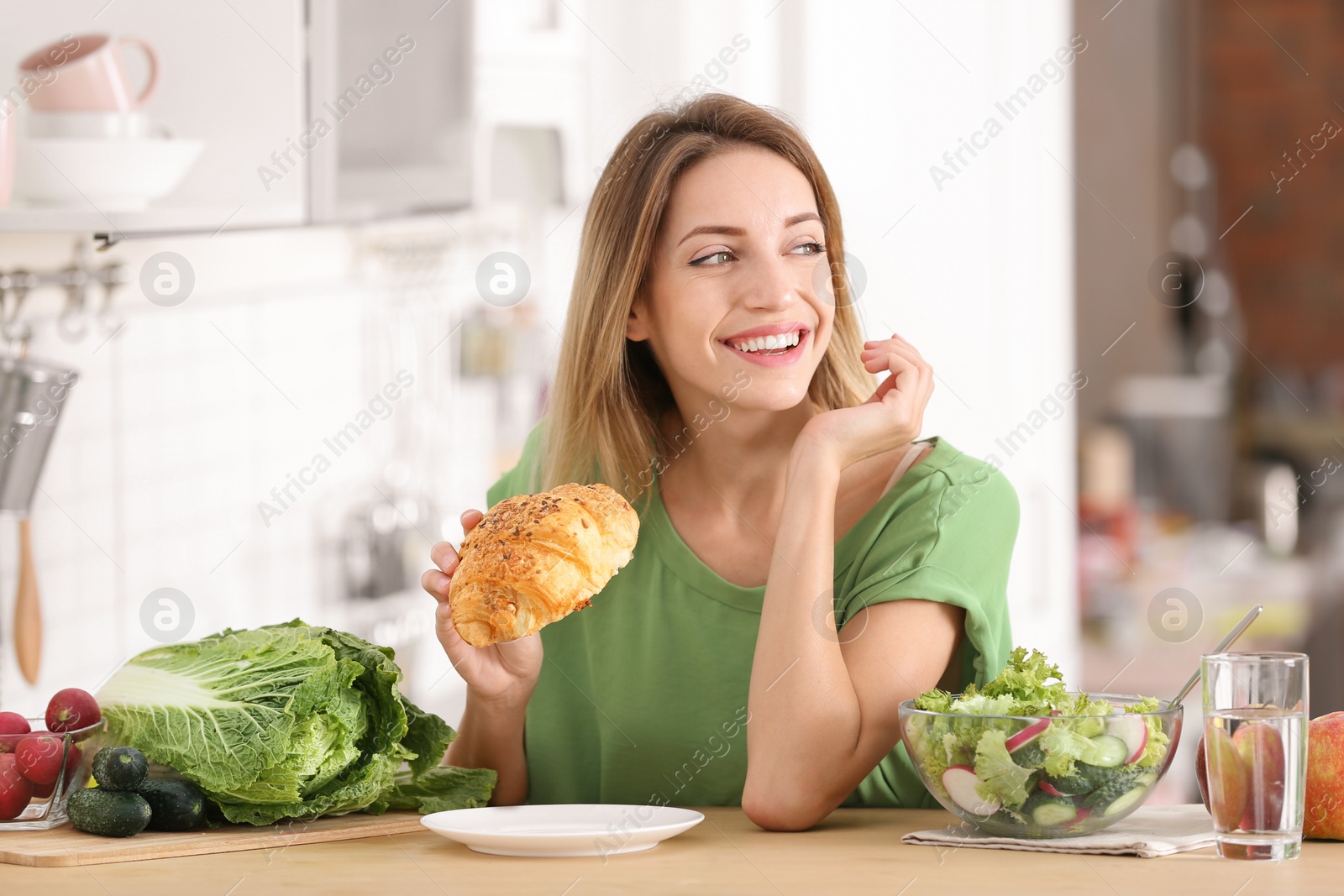 Photo of Woman eating croissant at table in kitchen. Diet failure