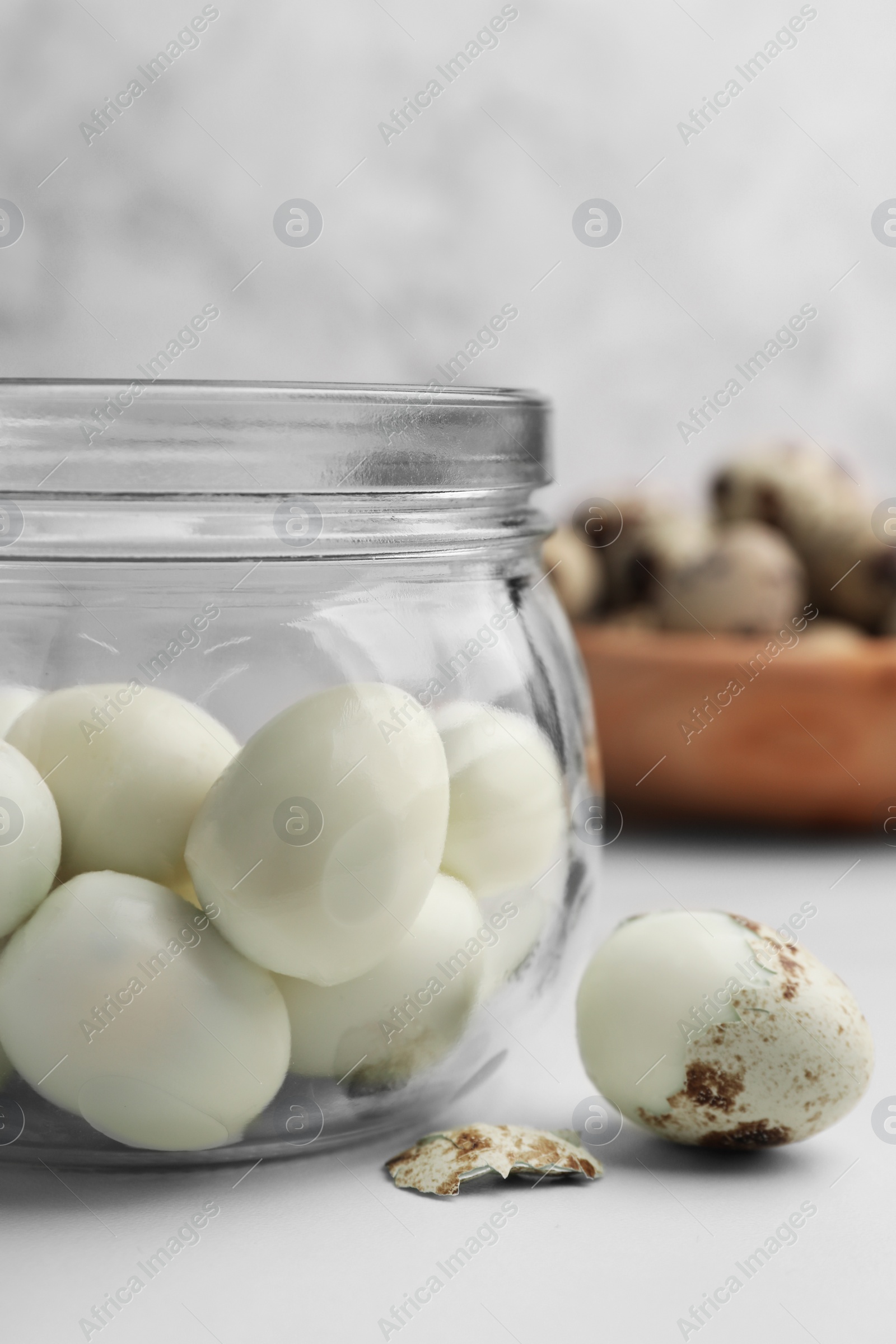 Photo of Glass jar with peeled boiled quail eggs and another one partly in shell on white table, closeup