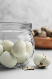 Photo of Glass jar with peeled boiled quail eggs and another one partly in shell on white table, closeup