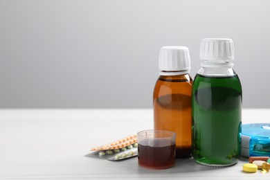 Bottles of syrup, measuring cup and pills on white table against light grey background, space for text. Cold medicine