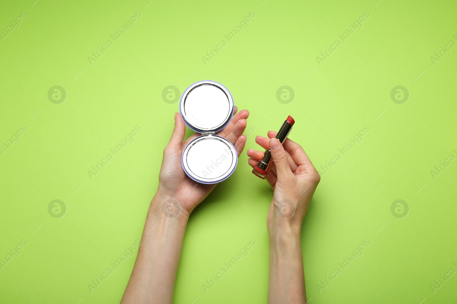 Photo of Woman holding pocket mirror and lipstick on light green background, top view