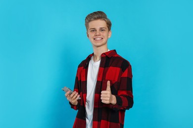 Photo of Teenage boy with smartphone showing thumb up on light blue background