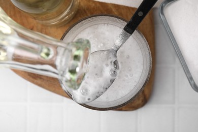 Photo of Pouring vinegar into spoon with baking soda over bowl at white tiled table, top view