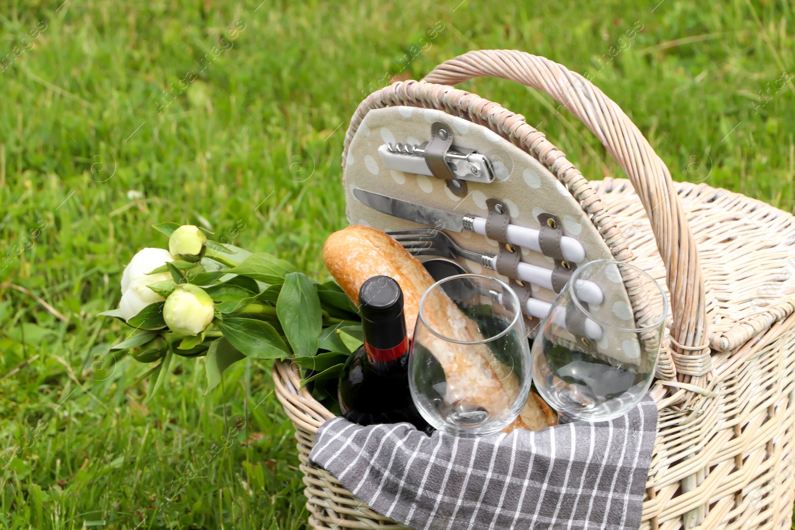 Photo of Picnic basket with wine, bread and flowers on green grass outdoors