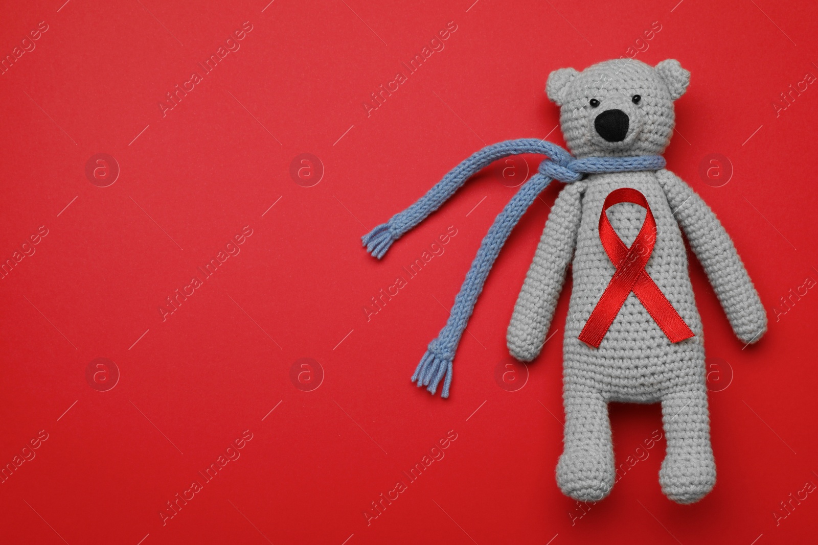 Photo of Cute knitted toy bear with ribbon on red background, top view and space for text. AIDS disease awareness