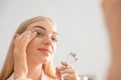 Photo of Attractive young woman curling her eyelashes indoors