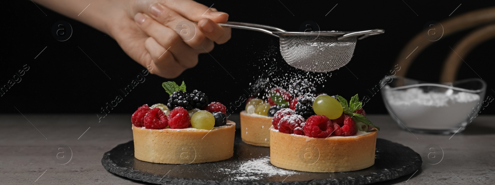 Image of Woman sprinkling delicious tartlets with powdered sugar at grey table, closeup. Banner design