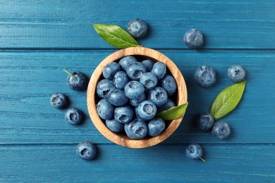 Photo of Bowl of fresh tasty blueberries with leaves on blue wooden table, flat lay