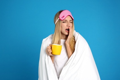 Young woman in sleeping mask wrapped with blanket holding cup on blue background