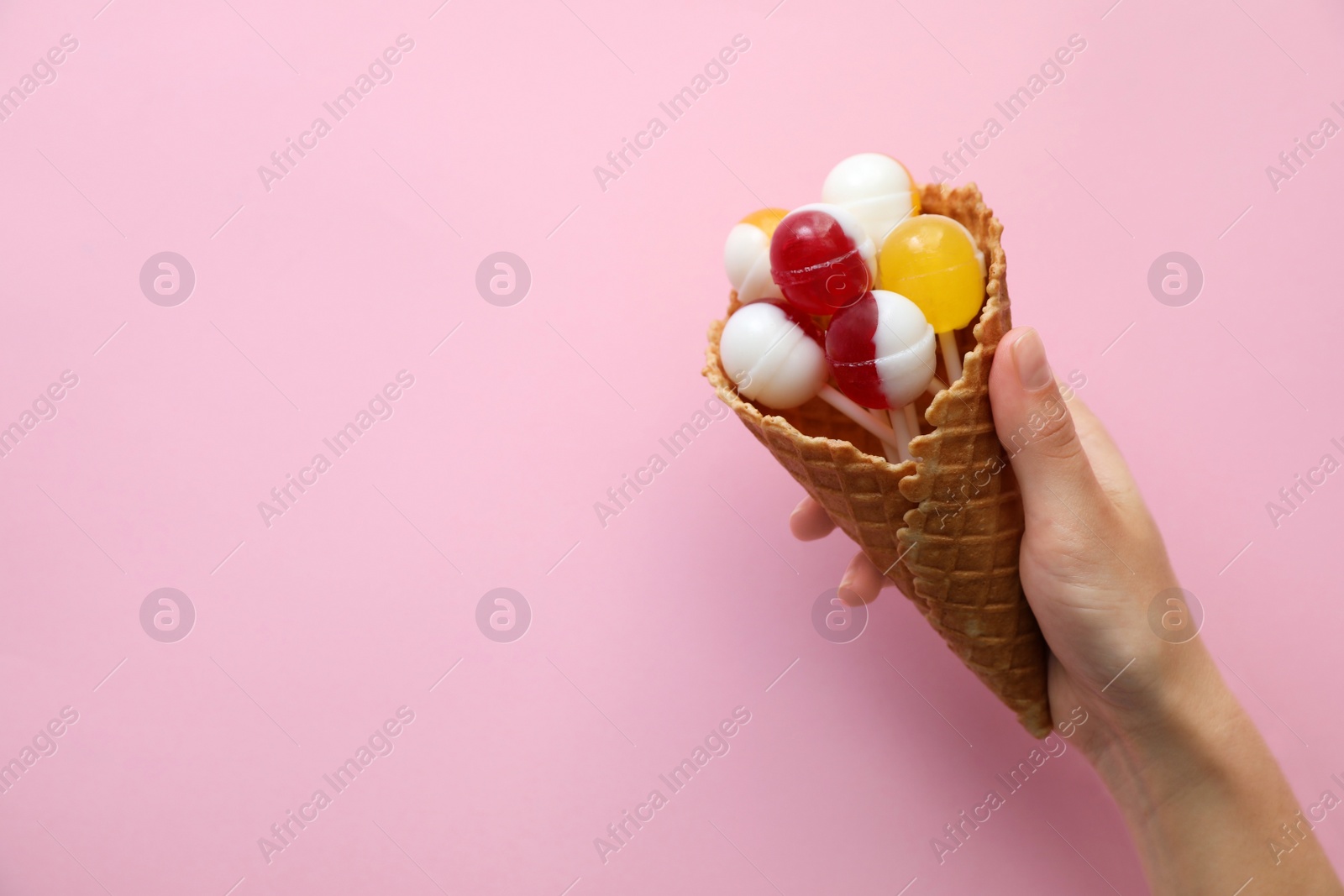 Photo of Woman holding wafer cone with delicious lollipop candies on pink background, top view. Space for text