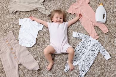 Little girl among baby clothes and detergent on carpet, top view