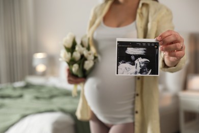 Photo of Young pregnant woman with flowers at home, focus on ultrasound picture of baby. Space for text