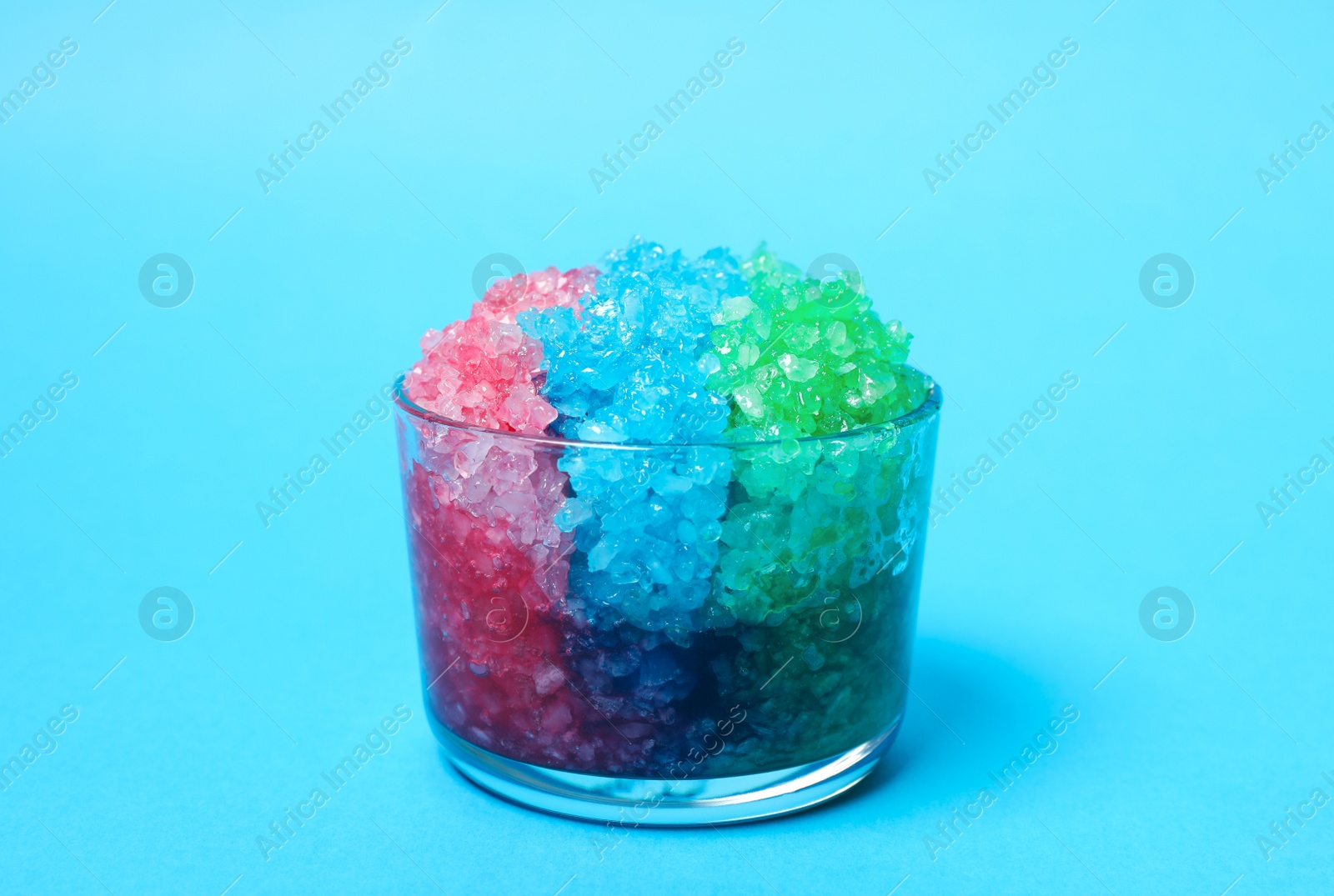 Photo of Rainbow shaving ice in glass dessert bowl on light blue background, closeup