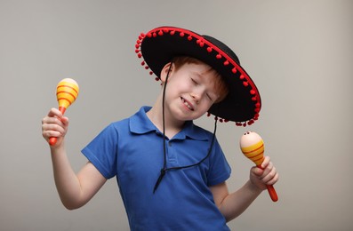 Cute boy in Mexican sombrero hat with maracas dancing on grey background