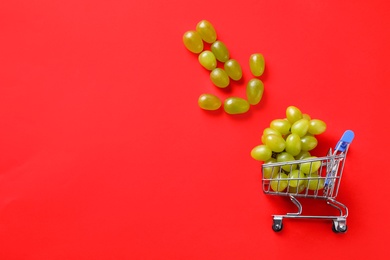 Arrow made of grapes pointing at small shopping cart with berries on red background, flat lay. Space for text