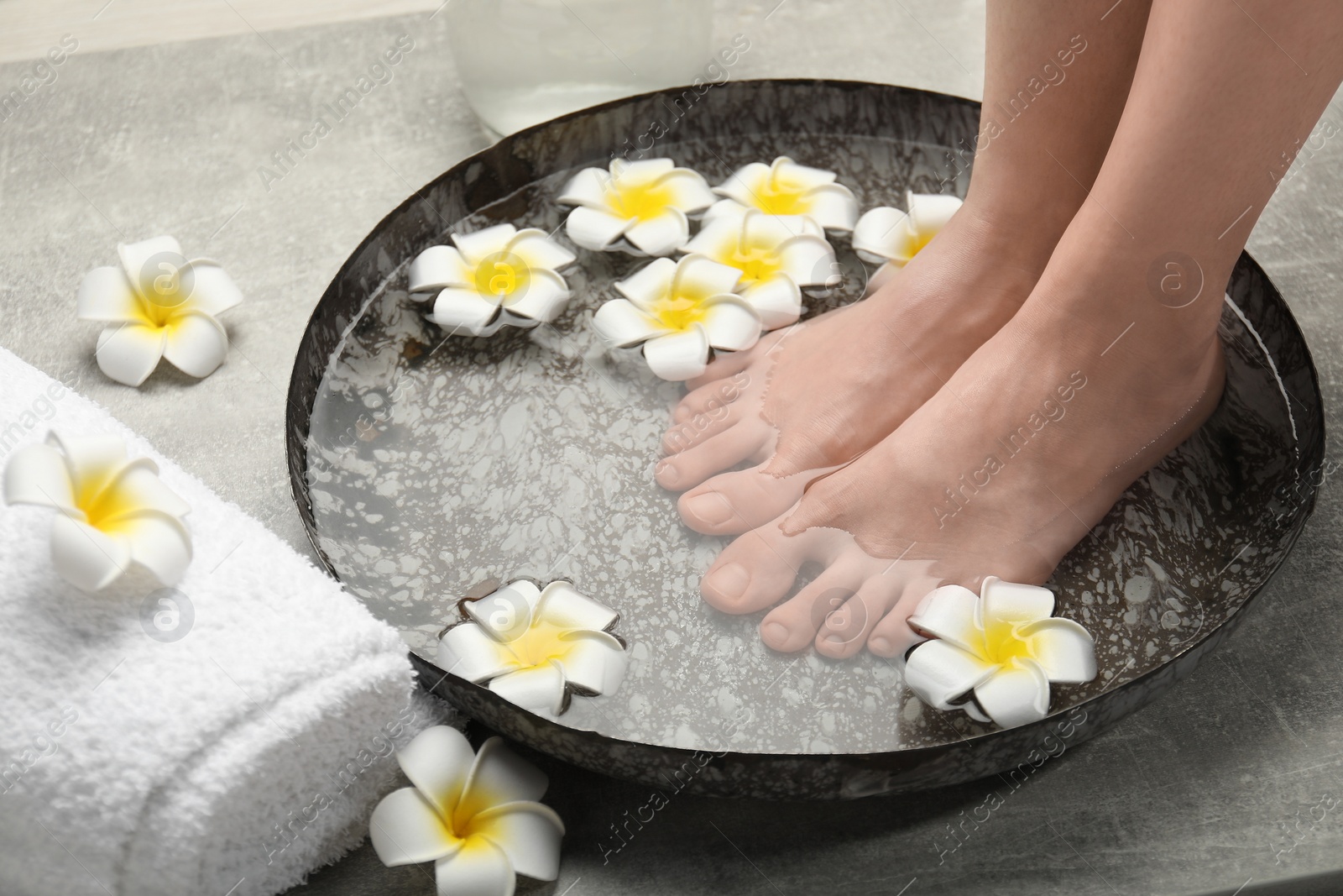 Photo of Woman soaking her feet in bowl with water and flowers on light grey floor, closeup. Spa treatment