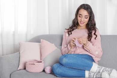 Photo of Young woman in warm sweater knitting on sofa at home. Space for text